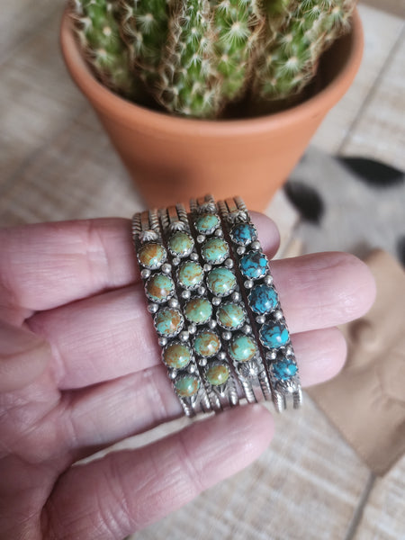 A sterling silver cuff bracelet featuring multiple Kingman turquoise stones, held in a person's hand with a cactus in the background.